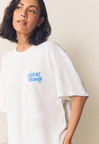 Camiseta blanca de algodón con un gráfico azul que dice "THE SUNDAY BRUNCH CLUB", con un corte relajado y mangas cortas.