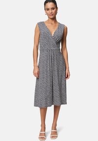 Sleeveless, navy dress with a deep V-neck and patterned texture in white shapes. Flared skirt falls to mid-calf. Paired with strappy white sandals.