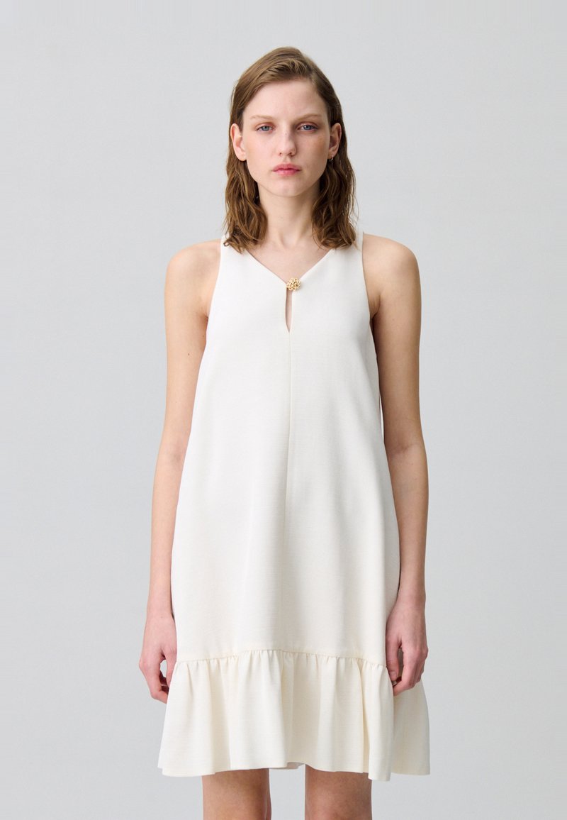 Vestido blanco sin mangas con escote en V y abertura en forma de llave, con un dobladillo con volantes y un detalle decorativo dorado en el escote.
