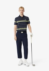 Polo a righe navy con righe verdi, abbinato a pantaloni navy. Il modello indossa scarpe da golf bianche e tiene un bastone da golf con un guanto.