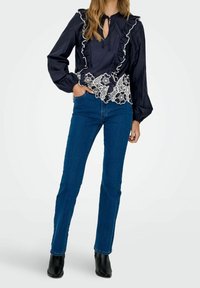 Femme portant un chemisier bleu marine avec une broderie florale en dentelle blanche et des volants, associé à un jean droit bleu et des bottines noires.