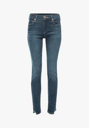 Osoba ubrana w ciemnoniebieskie obcisłe dżinsy z tylnymi kieszeniami, czarno-białą, pasiastą bluzkę z długim rękawem oraz czarne mokasyny, stojąca tyłem.