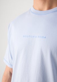 Světle modré bavlněné tričko s kulatým výstřihem a jemným logem "SCOTCH & SODA" tištěným v odpovídající modré barvě. Krátké rukávy a hladká textura.