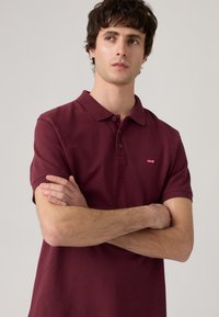 Burgundy színű pólóing rövid ujjakkal, bordázott gallérral, háromgombos légkörrel és kis Levi's logóval a mellkason. Simogató pamut textúra.