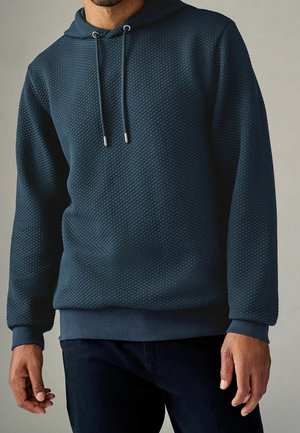 Homme portant un hoodie matelassé bleu foncé avec cordons et pantalon foncé assorti sur un fond uni.