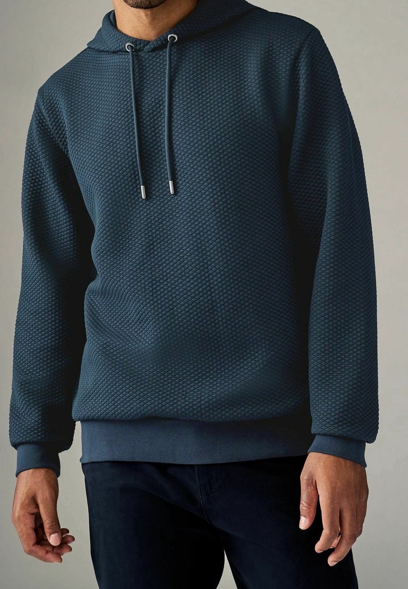 Homme portant un hoodie matelassé bleu foncé avec cordons et pantalon foncé assorti sur un fond uni.