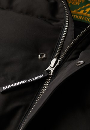 Giacca nera con zip argento, tirante con marchio "SUPERDRY EVEREST" e bottone a pressione circolare in metallo. Tessuto morbido e strutturato.