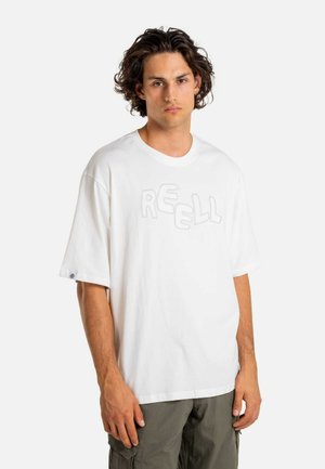 Young man with wavy hair wearing a loose white T-shirt with "REELL" text and olive green cargo pants, standing against a plain background.