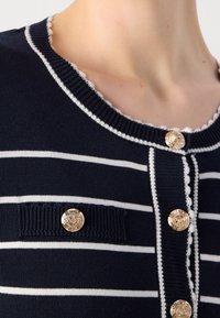 Marineblauwe gestreepte gebreide cardigan met witte strepen, voorzien van een geschulpte halslijn, gouden knopen en een voorzak met decoratieve knoop.