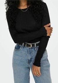 Schwarzes geripptes Langarmoberteil mit wellenförmigen Bündchen, getragen mit hochgeschnittenen hellblauen Jeans und einem schwarzen Gürtel mit goldener Schnalle.