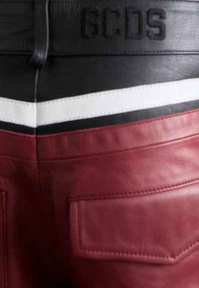 Pantalons en cuir rouge avec une taille noire ornée du logo « GCDS » en relief et des bandes blanches et noires autour de la taille. Deux poches arrière visibles.