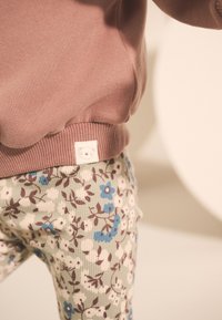 Roze sweatshirt met geribbelde zoom en een berenlogo-label; groene corduroy broek met bloemenprint in blauw en wit met bruine accenten.