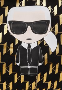 Figura de caricatura sobre fondo negro con patrones geométricos dorados. La figura lleva gafas de sol, traje negro, camisa blanca y corbata.