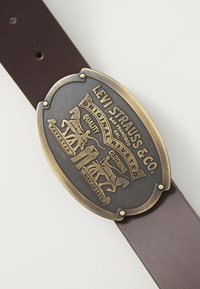 Brown leather belt featuring a large, oval, antique brass buckle with engraved text and horse graphics, highlighting brand origin and date.