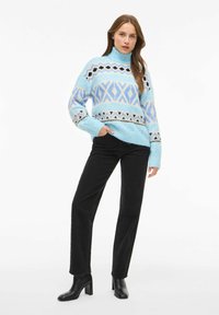 Hellblauer Strickpullover mit geometrischen Mustern in Blau, Schwarz und Creme. Kombiniert mit schwarzen hoch taillierten Hosen und schwarzen Stiefeln mit Absatz.