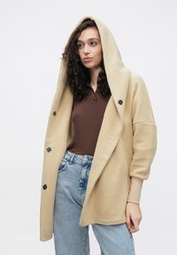 Casaco de lã bege com capuz grande, detalhes em botões e corte largo; usado por cima de uma blusa castanha canelada e jeans de ganga azul clara.