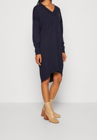 Robe marine à col en V avec des manches longues, coupe ample et ourlet asymétrique. Associée à des bottines à talons texturées beiges.