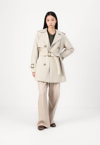 Beige trenchcoat med doble knapper, belted midje og skulderepauleter, kombinert med kremfargede bukser og sko med leopardmønstret.