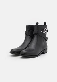 Une paire de bottines noires en cuir avec des talons bas, des sangles enroulées et des boucles en argent au logo MK sur les côtés, sur fond blanc uni.