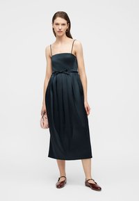 Γυναίκα που στέκεται με μια μαύρη midi φόρεμα χωρίς μανίκια με λεπτές τιράντες, κρατώντας μια μικρή ροζ τσάντα, φορώντας καφέ επίπεδα παπούτσια.