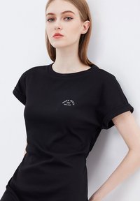 Camiseta negra hecha de algodón suave, con mangas enrolladas y un pequeño logo de la marca en blanco en el pecho. Textura suave y diseño ajustado.