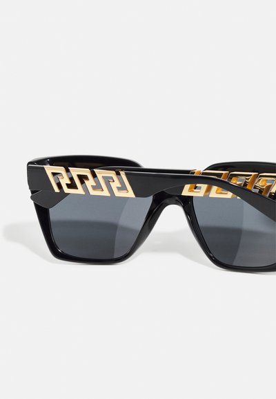 Óculos de sol pretos com um design geométrico, apresentando detalhes em dourado nas armações e lentes escuras. A forma ousada e angular enfatiza a modernidade.