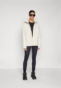 Jachetă bej ușoară cu glugă, combinată cu colanți negri mulați și încălțăminte neagră, purtate de o persoană cu ochelari de soare.