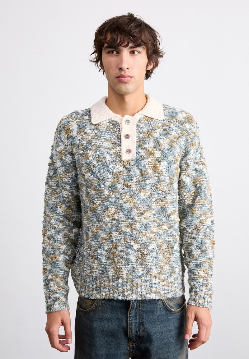 Maglione lavorato a maglia in tonalità blu, bianco e oro. Presenta un colletto color crema e una chiusura a tre bottoni. La texture sembra morbida e soffice.