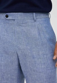 Pantaloni in lino azzurro chiaro con una texture sottile, dotati di chiusura a bottone nero e tasche laterali, mostrati dalla vita alla coscia.