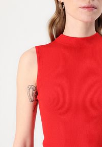Punainen ribbikuvioinen hihaton toppi, jossa on korkea pääntie ja teksturoitu kangas. Ylävarressa näkyy tatuointi.