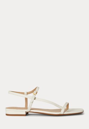 Sandale plate blanche avec fines lanières, boucle dorée et design à bout ouvert sur un fond clair.