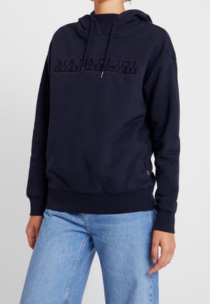 Osoba ubrana w granatową bluzę z kapturem z logo "Napapijri geographic" oraz jasnoniebieskie dżinsy, stojąca na tle jednolitego tła.