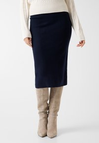 Jupe crayon en maille bleu marine, texture côtelée, longueur genou. Associée à des bottes à talons en daim taupe, au design élégant avec des boucles à la cheville.