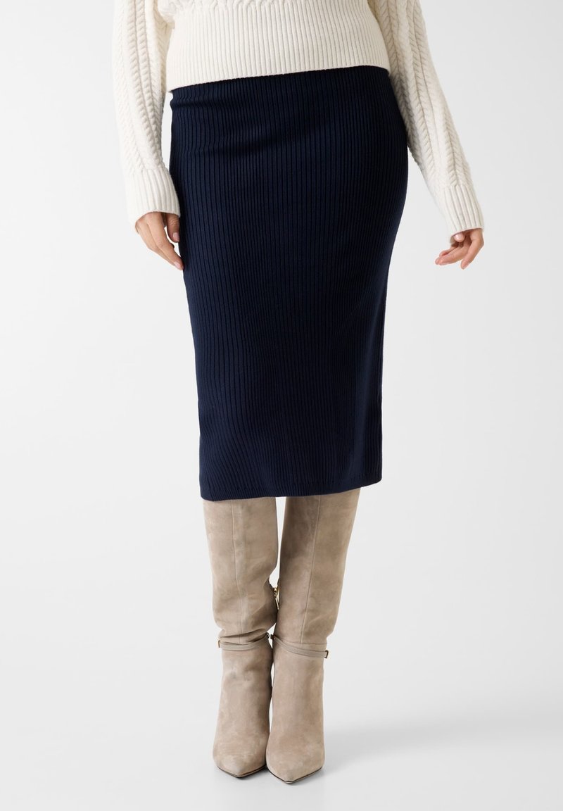 Jupe crayon en maille bleu marine, texture côtelée, longueur genou. Associée à des bottes à talons en daim taupe, au design élégant avec des boucles à la cheville.