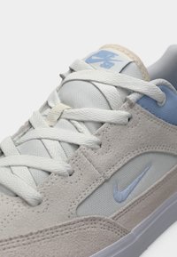 Nike SB kedas ar ādas un auduma augšdaļu gaiši pelēkā un zilā krāsā, ar baltām kurpju šņorēm un smalku zilu logotipu sānos.
