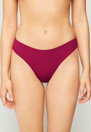 Kobieta w różowych, wysokich, prążkowanych majtkach bikini, widoczna od talii do połowy uda na jednolitym tle.
