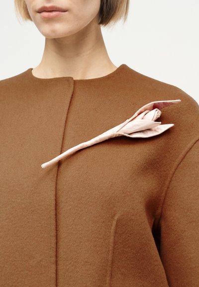 Femme portant un manteau marron avec une broche fleur en tissu rose pâle épinglée près de l'épaule sur un fond clair uni.