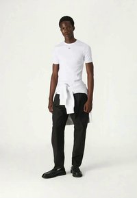 Homem vestido com uma camisa branca de mangas curtas justas, calças pretas, sapatos pretos e um suéter branco atado à cintura, de pé contra um fundo branco.