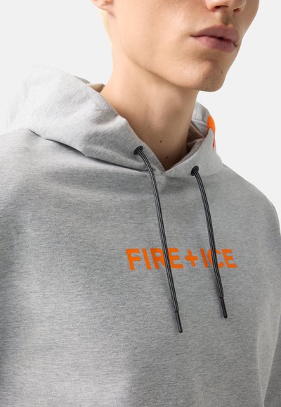 Gros plan sur une personne portant un sweat à capuche gris clair avec des cordons noirs et le texte orange "FIRE + ICE" sur la poitrine.
