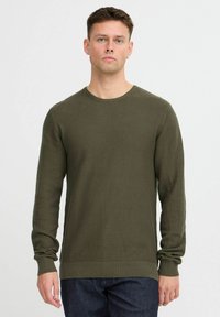 Olivengrüner strukturierter Pullover mit Rundhalsausschnitt, langen Ärmeln und gerippten Bündchen sowie Saum. Über dunklen Jeans getragen.