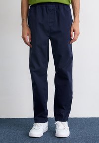 Pantaloni dintr-un amestec de bumbac de culoare bleumarin, cu o croială lejeră și talie elastică, asortați cu adidași albi. Textură fină, detalii minime.