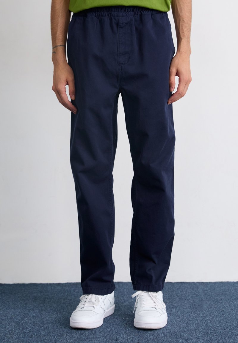 Pantaloni dintr-un amestec de bumbac de culoare bleumarin, cu o croială lejeră și talie elastică, asortați cu adidași albi. Textură fină, detalii minime.