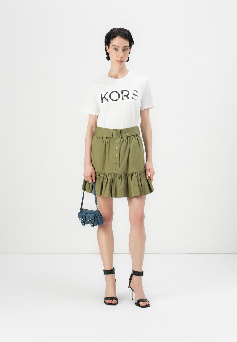 Maglietta bianca con logo "KORS", gonna svasata verde oliva con bottoni e cintura, sandali neri con tacco alto, mini borsa blu. Sfondo semplice.