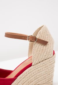 Sandale à talon compensé avec plateforme beige tissée, empeigne en tissu rouge et bride de cheville marron réglable avec boucle dorée.