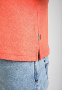 Koralna majica s kratkimi rokavi z teksturiranim geometrijskim vzorcem, z bočnim razporkom in majhno oznako blagovne znamke na robu. Oblečena z svetlo modrimi kavbojkami.