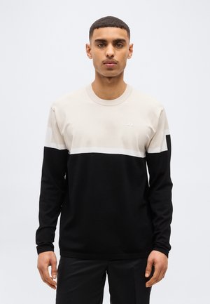 Giovane uomo con capelli corti che indossa un maglione a maniche lunghe a blocchi di colore nero e beige, in piedi davanti a uno sfondo chiaro e semplice.