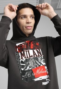 Felpa nera con cappuccio caratterizzata da una grafica frontale con "Forza Milan," "A.C. Milan" e un logo circolare. Il materiale sembra morbido con una finitura testurizzata.