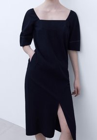 Robe bleu marine à encolure carrée, manches courtes bouffantes et fente latérale. Fabriquée en tissu texturé avec des détails de couture visibles.