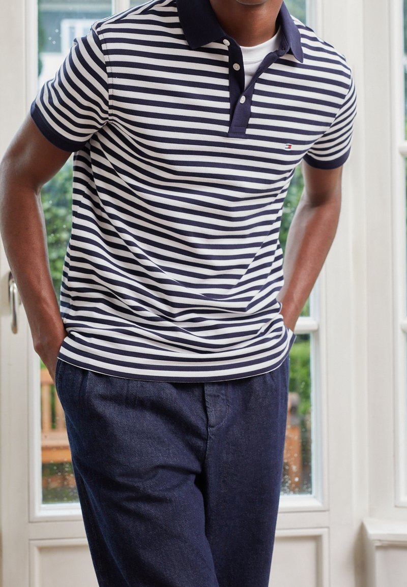 Polo rayé bleu marine et blanc avec un col et trois boutons, associé à un pantalon bleu marine foncé, se tenant à l'intérieur près d'une fenêtre.