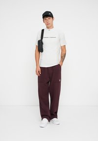 Ung mand iført en sort kasket, hvid Nike Tech t-shirt, maroon træningsbukser, hvide sneakers og en sort crossbody taske, stående mod en hvid væg.